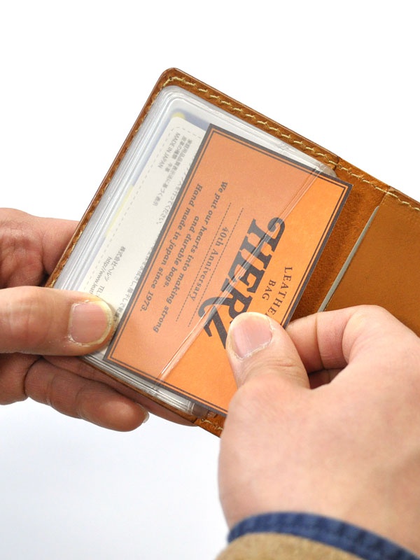 棕色皮革卡片收納夾，內有透明卡槽，展示一張印有黑色HERZ字樣的橘色卡片，雙手正在插入卡片。