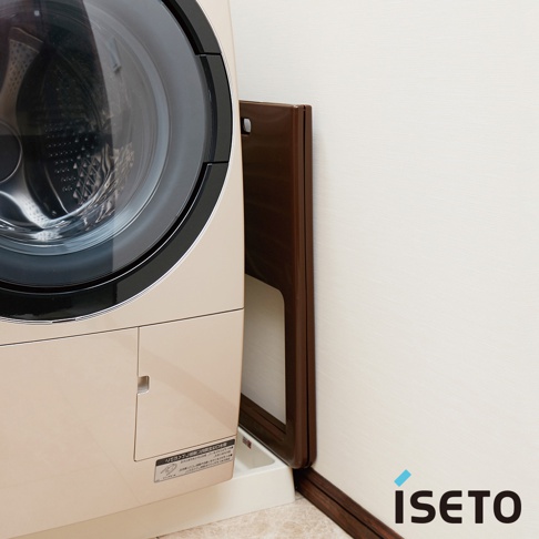 淺米色洗衣機旁，一個深棕色折疊式高腳置物籃靠牆立著，籃框為深棕色，籃板為淺白色。