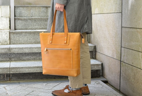 職人手作的淺棕色皮革威靈頓直式拉鍊托特包，包面有兩個外部拉鍊口袋，一個帶有 D 型環拉鍊頭，另一個是較小的方形口袋，以鉚釘固定。