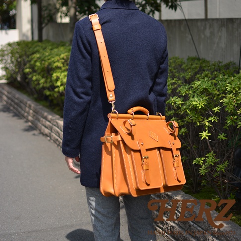 職人手作皮革旅行包 A-102，採用橘棕色皮革，設計有兩個前袋、頂部提把和可調式肩帶，搭配金屬扣環與縫線細節，適合肩背或斜背。