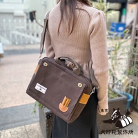 棕色純棉帆布製成的橫式側背包，配有皮革飾邊和提把，可肩背或手提。