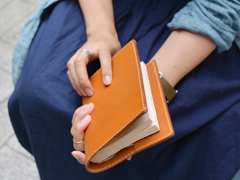 一只手拿著一本棕色皮革製的書套，裡面夾著一本白色筆記本。書套邊緣有縫線，整體呈現樸實的質感。