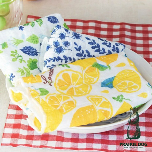 三條五層紗蚊帳質地擦拭巾，分別印有綠色植物、藍色花紋及黃色檸檬圖案，質地柔軟吸水，疊放在白色盤子裡，背景為紅色格紋桌布。