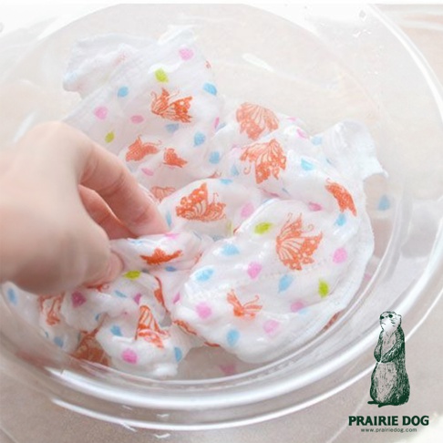 一塊白色五層紗質地的擦拭巾，上面印有橘色蝴蝶圖案及藍色、粉色、綠色圓點。一隻手正將擦拭巾浸入透明容器中。