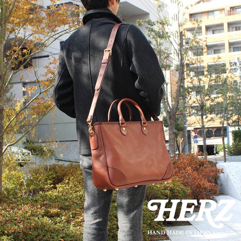 一位背著日本HERZ棕色皮革兩用商務托特包的男性，包款採用經典的職人手作設計，配有堅固的提把與可調整肩背帶，包身下方設有金屬腳釘，整體呈現沉穩優雅的商務休閒質感。