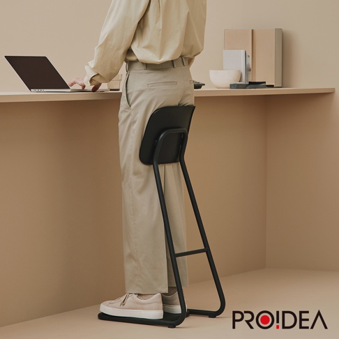 黑色STAN CHAIR人體工學站立椅，以金屬椅架搭配一體成形黑色椅面，椅面曲線貼合身體，黑色支架延伸至地面，支撐使用者站立。