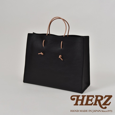 黑色牛皮手提包，配有兩條淺棕色皮革提把。包身設計簡約，正面有兩對皮革繫帶裝飾，可調節開口大小。