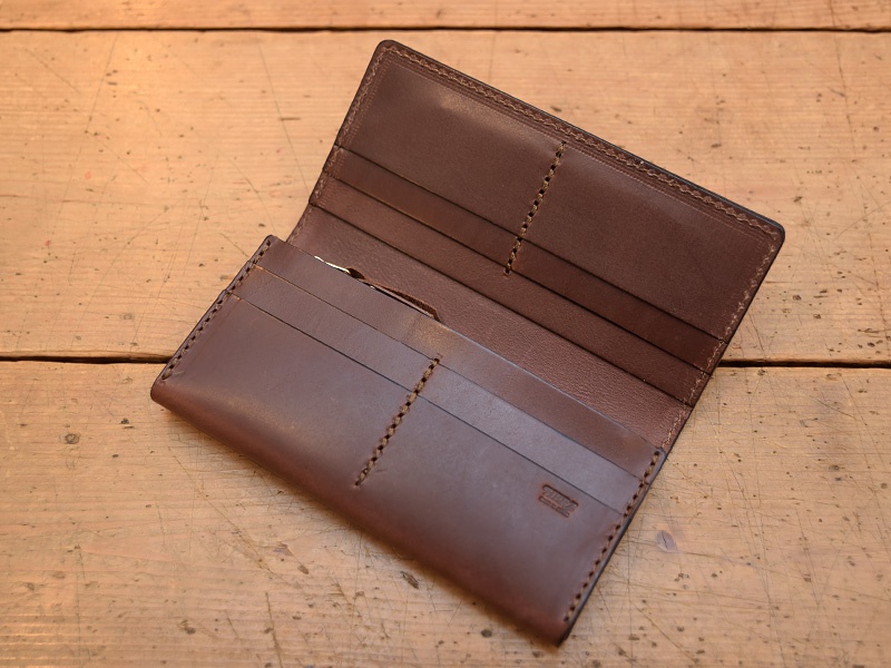 職人手作深棕色皮革長夾，打開狀態，內有多個卡片夾層，縫線精緻，皮革質感良好，置於木紋背景上。