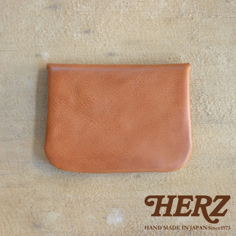 淺棕色皮革製成的職人手作小物收納袋，表面有自然皮革紋理，設有鈕扣式開合，整體呈現簡潔的方形設計，置於木紋背景上。