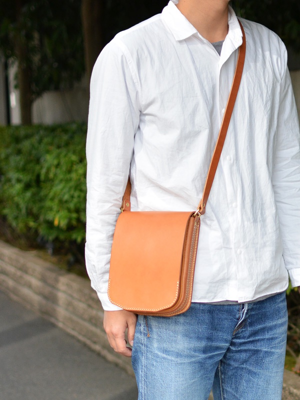 淺棕色皮革翻蓋直式單肩包，皮革表面光滑，邊緣有縫線裝飾，搭配棕色皮革肩帶。