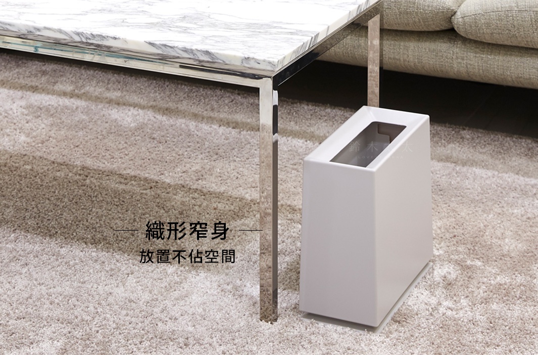 白色方形垃圾桶，傾斜開口，放置於淺色地毯上，旁邊為大理石桌面與金屬桌腳。