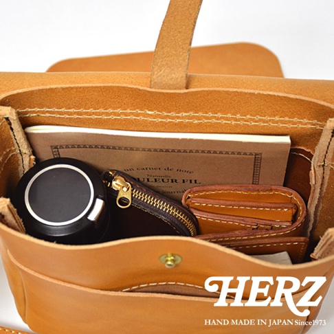 HERZ 品牌標誌和「HAND MADE IN JAPAN Since1973」字樣，下方為一個打開的棕褐色皮革斜背包，裡面裝著一本筆記本、一個黑色圓形小物、一個深棕色拉鍊零錢包以及一個棕褐色皮夾。