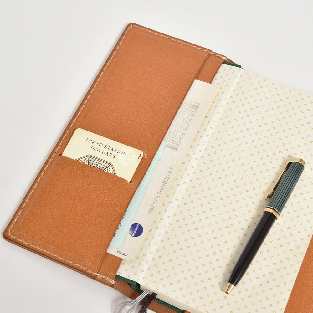 職人手作皮革旅行記事本書套，採用棕色皮革，內有卡槽，可放置卡片、票據。書套內頁為帶有金色圓點圖案的紙張，旁邊放置一支綠色與黑色搭配的鋼筆。