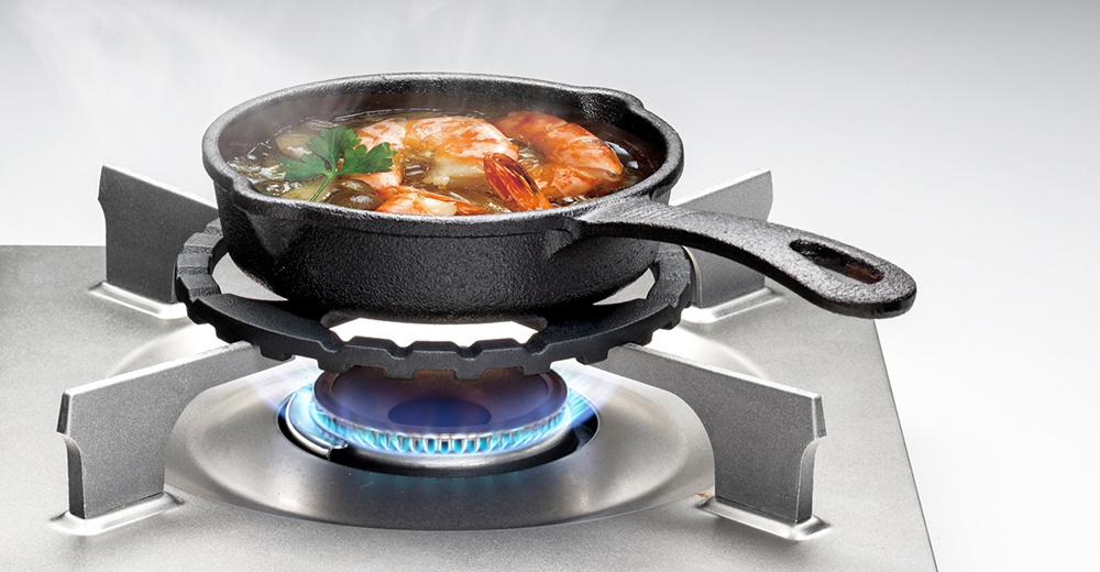 五德瓦斯爐架，黑色鑄鐵材質，圓形造型，帶有鋸齒狀邊緣，置於不鏽鋼瓦斯爐上，爐架上放著一個黑色鑄鐵小鍋，鍋內有蝦和香料，正在加熱中，散發出蒸氣。
