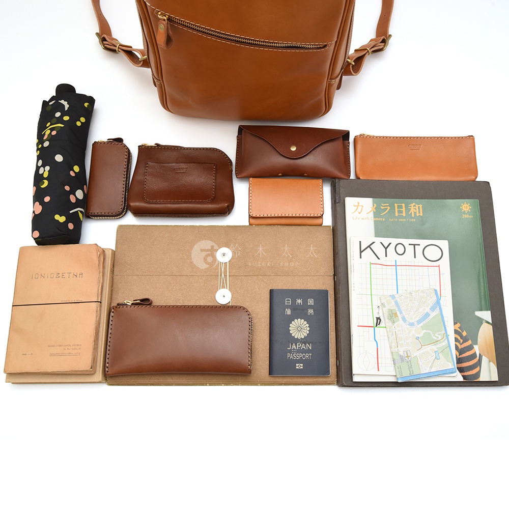 職人手作皮革長方型多口袋後背包，展示多樣配件，包含傘、皮夾、筆袋、筆記本、護照，以及京都旅遊指南等。