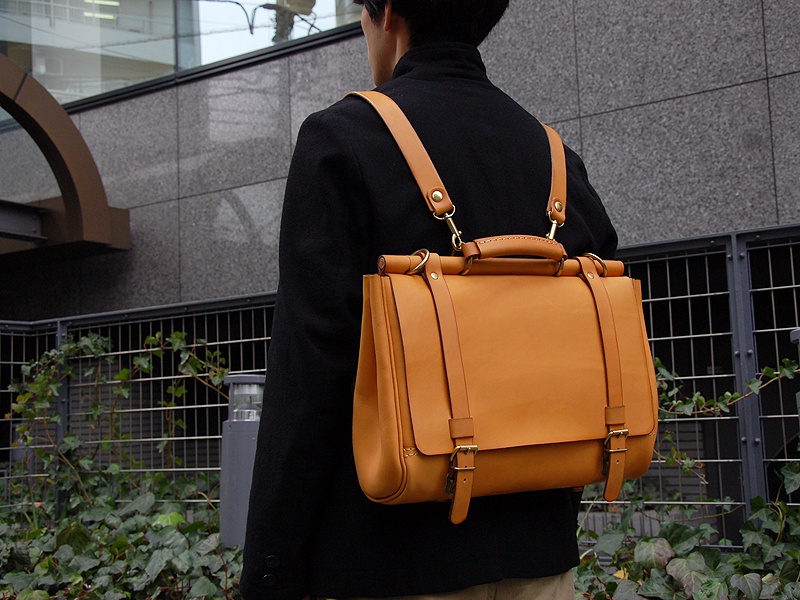 一款淺棕色皮革製成、屋瓦造型的三用側背包，具有雙邊金屬扣帶及頂部提把，背部配有可拆卸的皮革背帶。