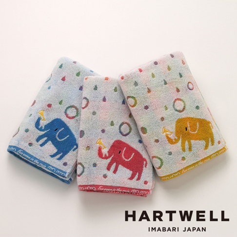 三條疊放的 Hartwell 今治毛巾，分別為淺藍、淺灰與淺黃色底，每一條毛巾上皆繡有一隻可愛的彩色小象，象鼻正吹著小號，毛巾邊緣印有 Fun even in the rain 字樣與雨滴、圓圈等圖案，整體風格童趣溫馨。
