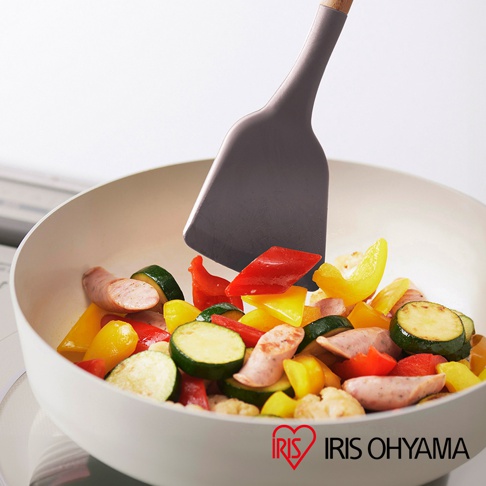 灰色矽膠鍋鏟正在翻炒鍋中切塊的香腸、櫛瓜、紅黃甜椒與花椰菜，鍋子為白色。