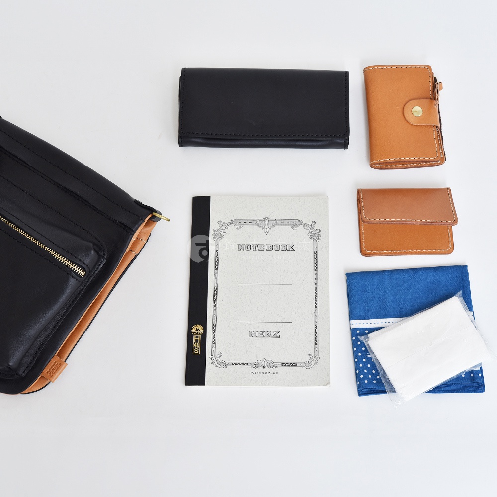黑色皮革翻轉斜背包，內裡為棕色皮革，搭配金色拉鍊。旁邊擺放著黑色長夾、兩款棕色皮革皮夾、一本封面為「NOTE BOOK」的筆記本，以及藍色小方巾與白色紙巾。