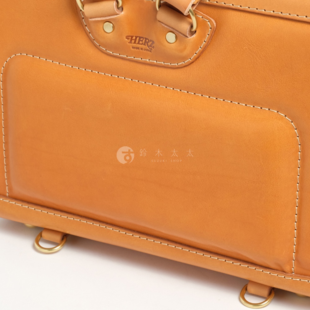 職人手作皮革橫式書包，特寫鏡頭展現天然皮革的溫潤色澤與細膩車縫線，包款上方的HERZ品牌標誌清晰可見，右側印有日文「鈴木太太」及英文SUZUKI SHOP字樣。