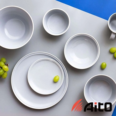 一組淺灰色美濃燒陶瓷餐具，包含不同尺寸的碗盤和杯子，擺放在灰色與藍色背景上，部分餐具裝飾有綠色葡萄。