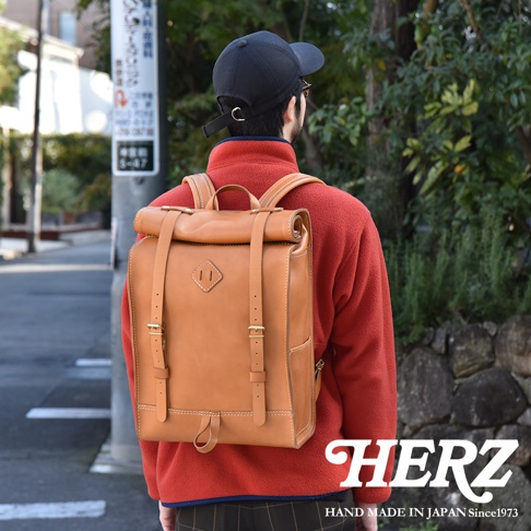 一個身穿紅色刷毛外套的人背著一個棕色皮革後背包，背包採用捲口設計，並配有可調節皮帶和金屬扣環。