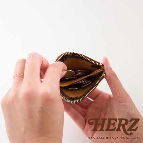 一雙手正打開一個日本HERZ職人手作棕色皮革零錢包，內部設有夾層並裝有少量硬幣，下方印有HERZ HAND MADE IN JAPAN Since1973字樣。