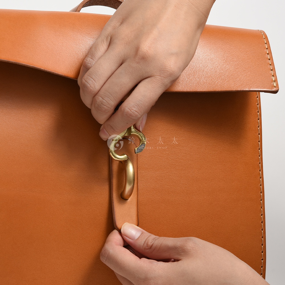 雙手正在操作一個棕褐色皮革後背包的鎖扣，鎖扣為金屬材質，設計簡潔。