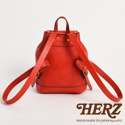 這是一款由日本品牌HERZ職人手工製作的紅色皮革迷你後背包，採用經典極簡設計，展示其皮革背帶與細緻縫線細節，背部印有HERZ品牌壓印標誌，下方並標註有品牌名稱與日本製造的標語。