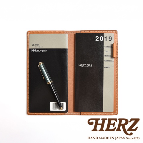 職人手作皮革輕便記事本書套，淺棕色皮革外殼，內含黑色筆記本和2019年曆。筆記本左側有「Handy pick」字樣，右側為年曆、月曆、個人備忘錄等分類標示。一支綠色與金色筆插在筆記本上。