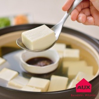 一只手拿著一個金屬湯匙，湯匙上盛著一塊白色的豆腐。湯匙下方是一個裝有醬汁的小白碗，置於一個裝滿湯水和豆腐塊的鍋中。
