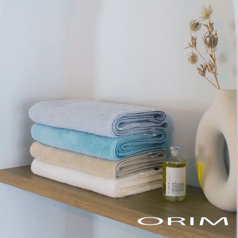 疊放的今治毛巾，由上至下顏色分別為淺藍、藍綠、米黃、白色，質地看起來柔軟吸水。旁邊有一個裝有透明液體的玻璃瓶和一個白色陶瓷花瓶，裡面插著乾燥的植物。