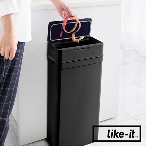 黑色長方形按壓式垃圾桶，桶蓋開啟，有人將蘋果皮丟入。