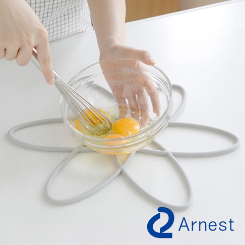 灰色矽膠鍋具防刮保護墊，呈花朵狀，上面放著一個裝有雞蛋的玻璃碗，有人正在用打蛋器攪拌。