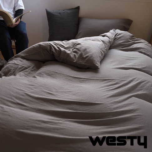 一張床鋪上鋪著灰色的被子，上面有幾張枕頭，其中一個枕頭是深灰色的，另一個是淺灰色的，被子上有明顯的褶皺，左側有一個人坐著，手中拿著一本書，圖片右下角有WESTY的文字標誌。