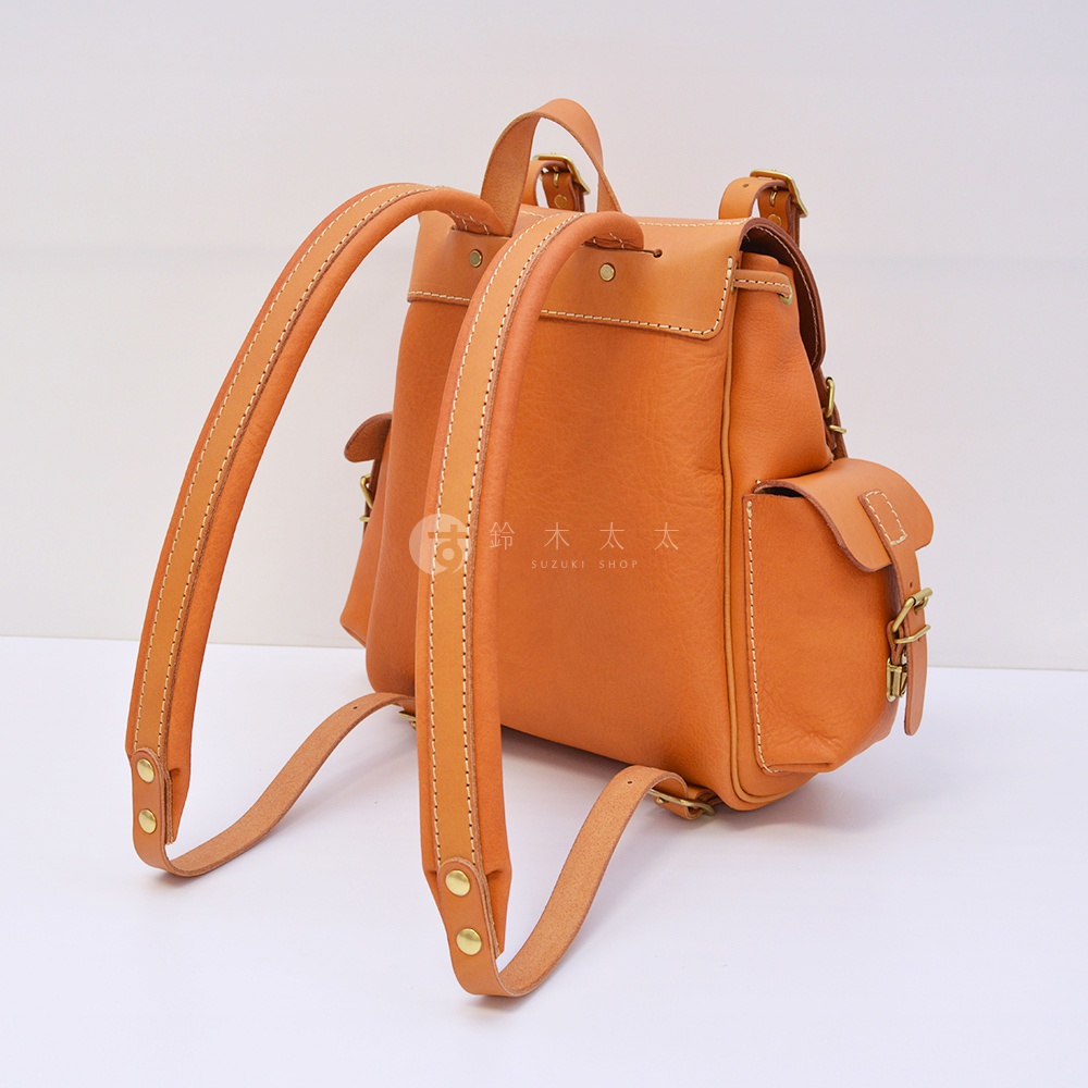 職人手作皮革復古風旅行後背包，採用天然皮革材質，呈現溫潤的焦糖橘色。包款設有兩個前袋，搭配金屬扣環及抽繩閉合，肩帶可調節，整體設計簡約實用。