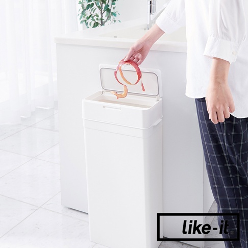 白色方形按壓式垃圾桶，配有透明蓋子，垃圾桶上部有一個按壓開啟的裝置，有人正在將果皮丟入桶內。