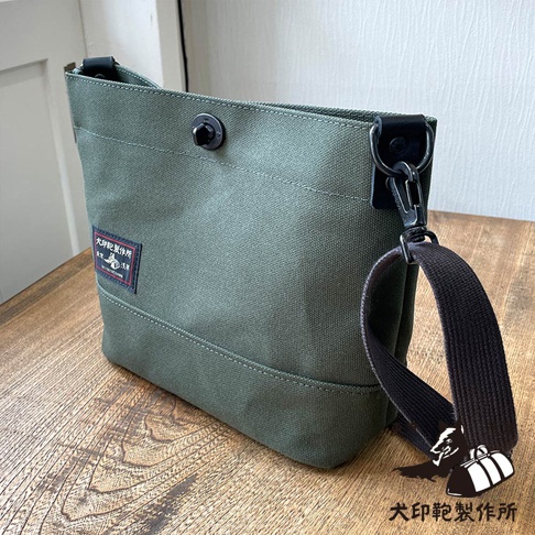 綠色帆布斜背包，帶有品牌標籤和可調節的黑色肩帶