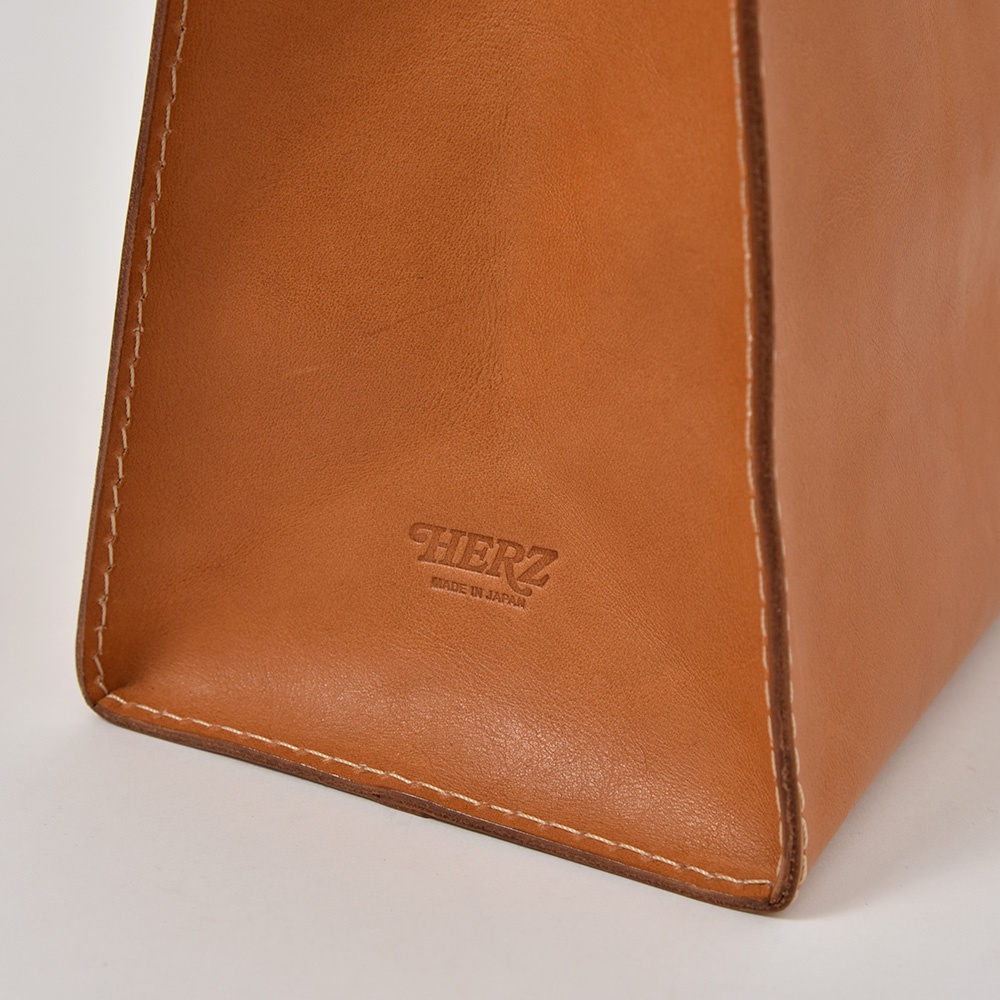 職人手作皮革提袋 T-5，採用天然皮革製成，呈現溫潤的棕褐色調。包款具有簡潔的線條與手工縫製的白色車縫線，質感細緻。皮革表面壓印有「HERZ」品牌標誌及「MADE IN JAPAN」字樣。