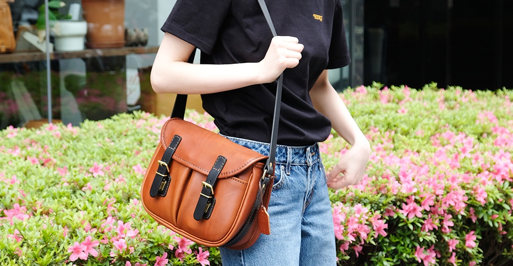 棕色皮革製成的傳統釣魚包，帶有黑色皮革飾邊和金屬扣，斜挎在穿著黑色T恤和牛仔褲的人身上。