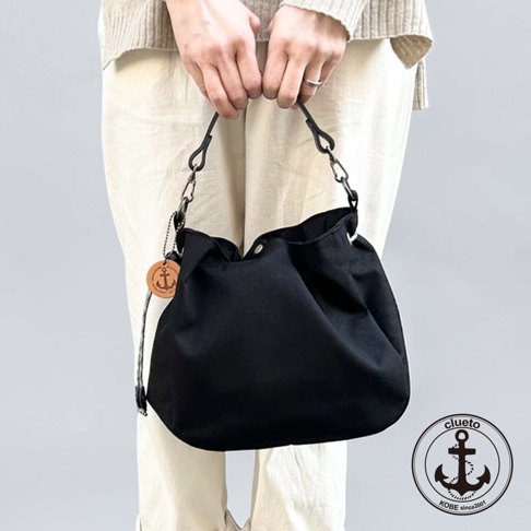 一位女性雙手提著一個神戶clueto品牌的黑色帆布防潑水手提包，包身圓潤柔軟，頂部設有按扣，側邊掛有品牌專屬的圓形錨圖案皮革吊飾，並搭配黑色金屬鉤扣與提帶。