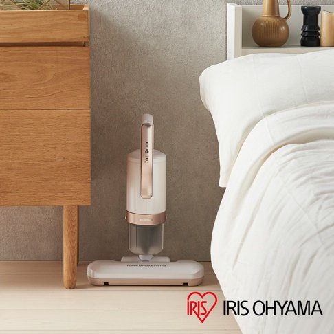 粉色機身、玫瑰金飾條的無線吸塵器，搭配同色系寬大吸頭，立於木質地板上，旁為白色床鋪與木製櫃子。