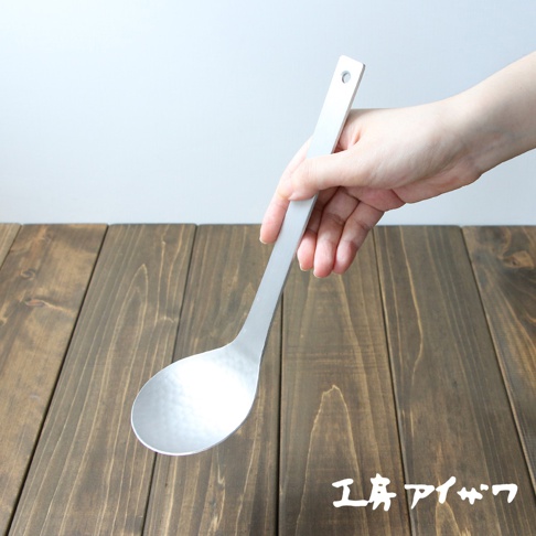 一隻手正拿著一個銀色鋁製湯勺，湯勺勺面有錘目紋，勺柄末端有一個小圓孔。