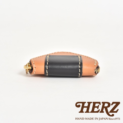 日本HERZ職人手作皮革三角飯糰造型零錢包，以淺棕色與黑色Latigo皮革拼接而成，側面展示拉鍊細節與縫線質感，右下角印有HERZ品牌標誌與產地資訊。