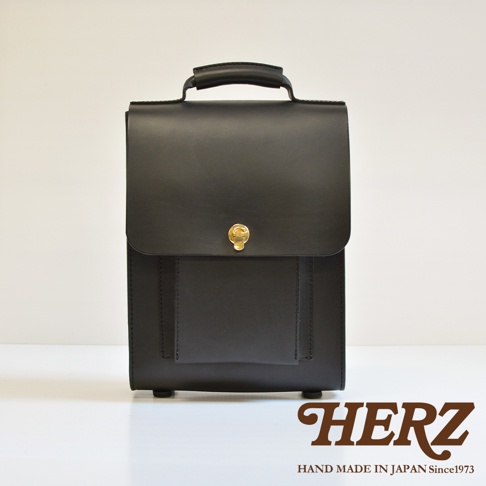 黑色皮革復古後背包，配有頂部提把、正面翻蓋設計及金色鎖扣，包身簡潔，下方有四個圓形腳釘。