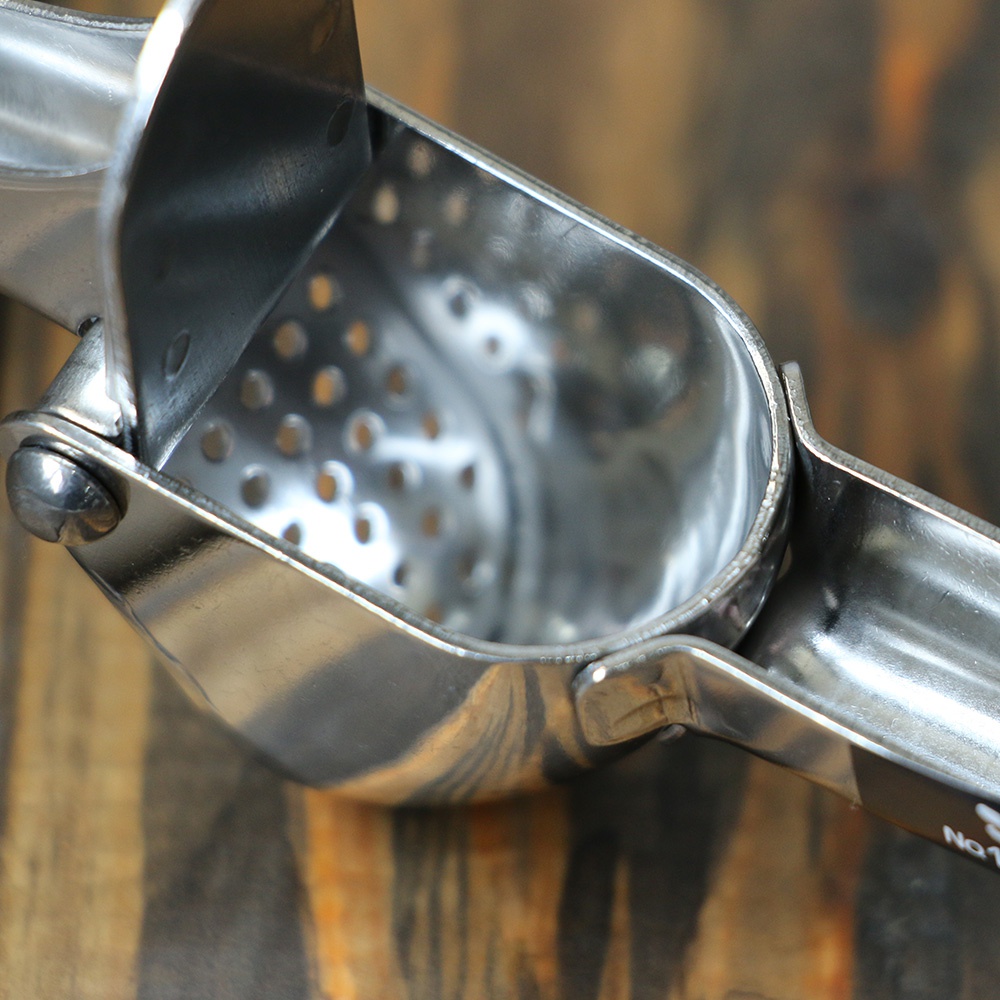 銀色不鏽鋼壓蒜器，帶有多個圓孔的蒜泥槽，置於木質背景上。