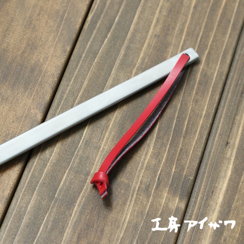 長條形銀色不鏽鋼鍋鏟，搭配一條紅色皮革掛繩，皮革掛繩末端打結，置於木紋背景上。