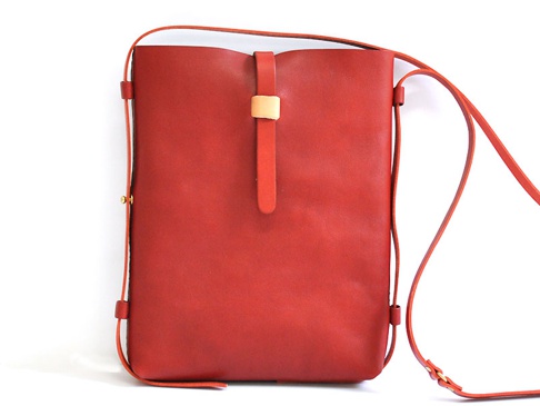 紅色皮革製成的直式單肩包，包身簡潔，正面中央有一條皮質束帶，束帶上附有淺棕色皮革裝飾，肩帶為細長皮質，兩側有金屬扣件。