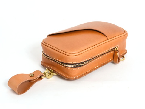 職人手作的淺棕色皮革斜背包，採用直立式設計，設有拉鍊開合及金屬扣環，搭配皮革掛繩。