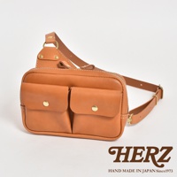 職人手作皮革雙口袋斜背包，採用柔軟的淺棕色皮革，設有兩個帶有金色金屬釦的方形前袋，以及一個拉鍊主隔層。包包配有可調節的皮革肩帶。